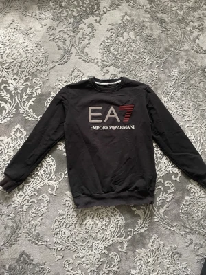 EA7 tröja  - Snygg svart sweatshirt från Emporio Armani med stort EA7-tryck i vitt och rött på bröstet. Tröjan har rund halsringning, långa ärmar och ribbade muddar vid ärmslut och nederkant. Perfekt för en trendig och avslappnad stil.