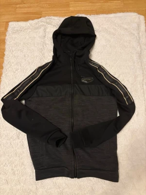 Svart och mörkgrå hoodie med dragkedja - Snygg hoodie från Supply & Demand i svart och mörkgrått med dragkedja framtill. Huva med dragsko och dekorativa ränder längs ärmarna samt Kappa-logga på bröstet. Tillverkad i mjukt och bekvämt material, perfekt för en avslappnad streetwear-look.