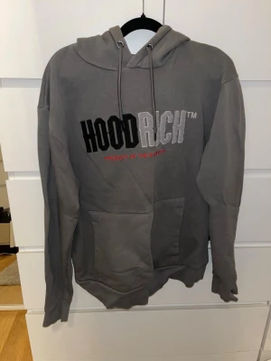 Grå HOODRICH hoodie med brodyr - Snörerna är slitna men annars är den bra. Original pris var 800kr. Snygg grå hoodie från HOODRICH med stor broderad logga på bröstet i svart, vitt och rött. Klassisk huva med dragsnören och stor magficka framtill. Perfekt för dig som gillar streetwear och vill ha något bekvämt och stilrent.