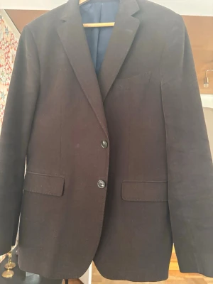 Gant- Mörkbrun kraftig kavaj i bomull - Snygg stilren mörkbrun kavaj med två knappar framtill och klassisk krage. Kavajen har två fickor med lock och en bröstficka. Tillverkad i ett mjukt tyg, helfodrad med mörkblått foder. Perfekt för en dressad men avslappnad look. 