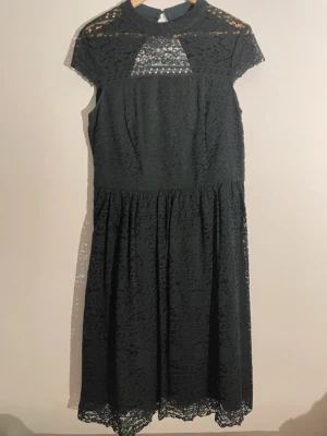 Svart spetsklänning med hög krage - Elegant mörkgrön klänning i spets med hög krage och korta ärmar. Klänningen har vackra spetsdetaljer över axlar och bröst samt en öppen rygg med dragkedja bak. Kjolen är vadlång och har en lätt volangkant nertill. Perfekt för dig som gillar en klassisk och stilren look.