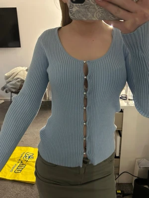 Ljusblå ribbad kofta med pärlknappar - Trendig ljusblå ribbad kofta med långa ärmar och djup rundringning. Koftan har dekorativa pärlknappar hela vägen framtill och är figurnära i modellen. Perfekt att styla med både jeans och kjol för en snygg look.