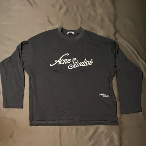 Mörkgrå longsleeve från Acne Studios - Snygg grå långärmad tröja från Acne Studios med vit logga tryckt framtill och mindre logga nere vid fållen. Tröjan har en avslappnad passform och är gjord i mjukt bomullsmaterial. Perfekt för en stilren och enkel look. Priset är ej hugget i sten så skäms it vid förslag🙂