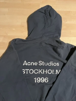 Mörkblå hoodie Acne Studios - Snygg mörkblå hoodie från Acne Studios med stor huva och tryck på ryggen där det står 'Acne Studios STOCKHOLM 1996' i vitt. Hoodien har en avslappnad passform och är tillverkad i mjukt bomullsmaterial. Perfekt för en cool och casual look. Storlek M men sitter väldigt baggy