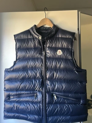 Mörkblå dunväst från Moncler - Snygg mörkblå dunväst från Moncler med glansig finish och klassisk logga på bröstet. Västen har två dragkedjeförsedda fickor framtill och hög krage. Perfekt för lager-på-lager och riktigt trendig look. Storlek 3