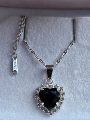 Halsband med svart hjärthänge - Snyggt halsband i rostfritt stål med ett hjärtformat svart hänge omgivet av gnistrande klara stenar. Kedjan har en tvinnad design och justerbar längd. Perfekt för dig som gillar statement-smycken med elegant touch.