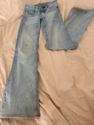 Ljusblåa bootcut jeans Cheap Monday weekday - Säljer ett par ljusblå bootcut jeans från weekday med cool grön patch och tryck bak i midjan. Jeansen har klassisk femficksdesign. Har en liten mini fläck på högra knäet.