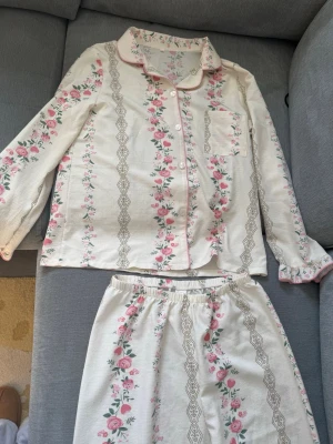 Blommig pyjamas med spetsdetaljer - Söt tvådelad pyjamas i cremevitt tyg med rosa blommönster och dekorativa spetsinspirerade ränder. Skjortan har krage, bröstficka och knappar framtill, medan byxorna har resår i midjan. Perfekt för mysiga kvällar hemma.