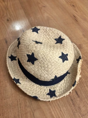 Stråhatt med blå stjärnor - Cool stråhatt med mörkblå stjärnor och matchande blått band runt hatten. Hatten har en klassisk form och är perfekt för soliga dagar. Tillverkad i flätad strå och har en ljus beige färg som kontrasterar snyggt mot de blå detaljerna.