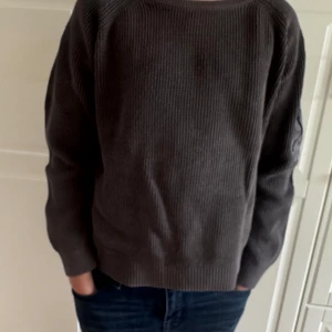 Fet grå stickad tröja  - Mörkgrå stickad tröja i grov struktur med lång ärm och en cool ficka med dragkedja på ena ärmen. Tröjan har en avslappnad passform och ribbade muddar vid ärmslut och nederkant. Perfekt för dig som gillar en enkel men ändå unik stil.
