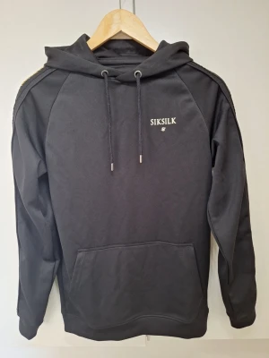 Svart hoodie från SikSilk - Svart hoodie från SikSilk med broderad logga på bröstet. Klassisk huva med dragsnören och känguruficka framtill.