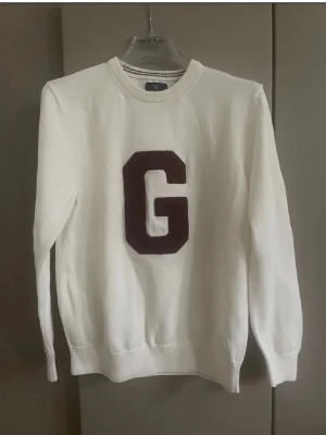 Vit stickad tröja med G-applikation i vinrött färg - Vit stickad tröja med en stor mörkröd G-applikation på bröstet. Tröjan har rund halsringning, långa ärmar och ribbade muddar vid ärmslut och nederkant. Perfekt för dig som gillar college-stil och enkel design. Storlek som på sista bilden men passar S
