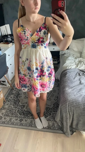 Blommig sommarklänning med v-ringning - Söt kort blommig klänning med v-ringad överdel och tunna axelband. Klänningen har en vit bas med färgglada blommor i rosa, blått, gult och lila. Tillverkad i ett lätt och luftigt material, perfekt för varma dagar. Passformen är normal och modellen är ärmlös.