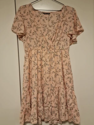 Ljusrosa blommig kortklänning - Söt ljusrosa kortklänning från SHEIN med blommigt mönster och volangdetaljer. Klänningen har omlott framtill, korta ärmar och en avslappnad passform. Perfekt för varma dagar och somriga tillfällen.