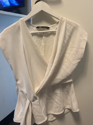 Vit omlottblus från Zara - Stilren vit omlottblus från Zara med knytning i sidan och mjukt, lätt tyg. Blusen har v-ringning och korta ärmar, vilket ger en avslappnad men chic look. Perfekt att matcha med jeans eller kjol för en trendig outfit. Även litet hål vid sidan där man knyter därav säljer jag för billigare pris.