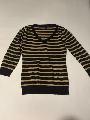 Randig stickad tröja med V-ringning - Snygg stickad tröja med svarta och beige horisontella ränder. Tröjan har V-ringning, kort ärmar och ribbade muddar vid ärmslut och nederkant. 