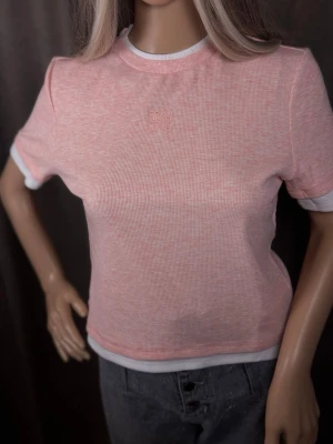 Rosa ribbad t-shirt från Miu Miu - Säljer en ljusrosa ribbad t-shirt från Miu Miu med broderad logga framtill. T-shirten har vita kontrastkanter vid ärmslut och halsringning samt en croppad passform. Perfekt för dig som gillar en söt och trendig stil.
