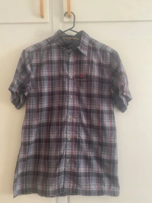 Rutig kortärmad skjorta i bomull - Kortärmad skjorta med rutigt mönster i blått, grått, vitt och röda detaljer. Klassisk krage och knäppning framtill. Tillverkad i lätt och sval bomull, perfekt för varmare dagar. Bröstficka med liten broderad detalj.