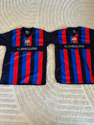Två FC Barcelona matchtröjor blå/röd S-M - Säljer två klassiska FC Barcelona matchtröjor i storlek S och M. Tröjorna har vertikala blå och röda ränder, klubbmärke på bröstet och texten 'FC BARCELONA' framtill. Tillverkad i ett lätt och snabbtorkande material, perfekt för fotboll eller träning.