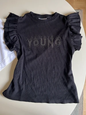 Svart topp med volangärmar Gina Tricot - Svart ribbad topp från Gina Tricot med volangärmar och texten 'YOUNG' i glittriga stenar framtill. T-shirten har en rund halsringning och är tillverkad i mjukt material som sitter snyggt på kroppen.
