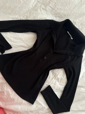 Svart ribbad långärmad topp - Stilren svart skjorta med ribbad struktur och lång ärm. Skjortan har klassisk krage och knappar framtill. Perfekt för dig som gillar en enkel men snygg look.