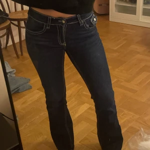 Mörkblå bootcut jeans med kontrastsömmar - Säljer ett par mörkblå bootcut jeans med vita kontrastsömmar och klassiska fem fickor. Jeansen har låg midja och en snygg passform. Perfekta för dig som gillar en tidlös och trendig look. Från Gina 
