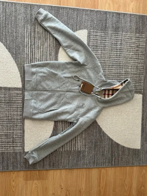 Grå hoodie från Burberry med dragkedja - Snygg grå hoodie från Burberry med dragkedja och klassiskt rutigt foder i huvan. Tröjan har två fickor fram, ribbade muddar och en stilren look. Perfekt för dig som gillar exklusiva detaljer och tidlös design. Materialet känns mjukt och bekvämt.