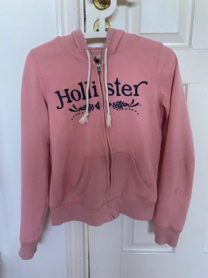Rosa hoodie från Hollister - Säljer en rosa hoodie från Hollister med dragkedja och fickor framtill. Stor logga broderad i mörkblått över bröstet och dekorativa snören i huvan. Mjuk bomullsmix och ribbade muddar vid ärmslut och nederkant. Perfekt för en chill och avslappnad stil. Dragkedjan kan var lite trög i början men funkar såjlary lika vanligt som andra, på tröjan går även några trådar lossnat från hollis tre t märket men det ser man på bilden. 