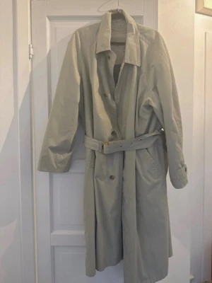 Beige trenchcoat med bälte - Stilren beige trenchcoat med klassisk krage och knäppning framtill. Jackan har ett matchande bälte i midjan och snedställda fickor. Materialet ser ut att vara bomull eller en bomullsblandning, perfekt för lager på lager. Enkel och tidlös design som passar till många outfits.