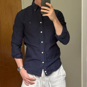 Mörkblå skjorta från Morris - Snygg mörkblå skjorta från Morris med klassisk krage och vita knappar. Skjortan har lång ärm och är tillverkad i mjuk bomull, perfekt för en clean och stilren look. Passformen är normal och den har diskret logga broderad på bröstet.