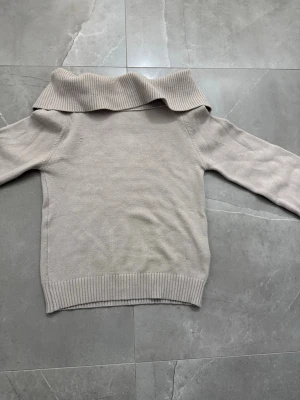 Beige stickad tröja med stor krage - Säljer en beige stickad tröja med lång ärm och bred, vikt krage. Tröjan har ribbade muddar vid ärmslut och nederkant, vilket ger en snygg och avslappnad look. Perfekt för dig som gillar stilrena och enkla plagg.