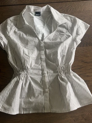 Randig kortärmad blus från Gina Tricot - Snygg vit blus med svarta vertikala ränder från Gina Tricot. Modellen har korta ärmar, markerad midja med resår och knappar framtill. Klassisk krage och lätt figursydd passform ger en fräsch och stilren look. Helt ny