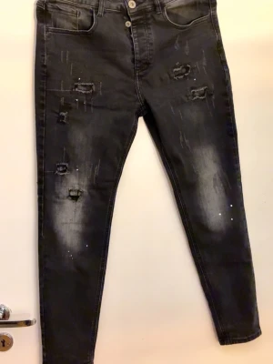 Svarta slitna skinny jeans - Säljer ett par svarta skinny jeans med slitningar och distressed detaljer på benen. Jeansen har hög midja, två knappar i gylfen och klassiska fem fickor. Materialet är stretchigt denim som sitter tight och bekvämt.