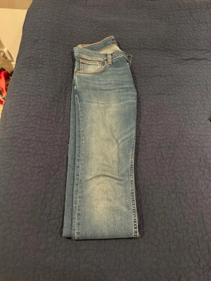 Nudie jeans Blå raka jeans med ljus tvätt - Säljer ett par klassiska blå jeans med rak passform och snygg ljus tvätt på låren. Jeansen har fem fickor, bälteshällor och kontrastsömmar. Perfekta till en avslappnad stil och funkar året runt.