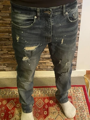 Mörkblå slitna jeans med hål - Säljer ett par mörkblå jeans med slitningar, hål och färgstänk för en riktigt trendig och avslappnad look. Jeansen har rak passform och klassisk femficksdesign. Perfekta för dig som gillar streetwear och en edgy stil.
