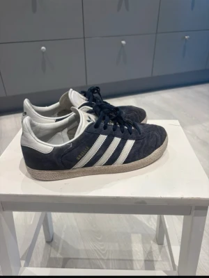 Adidas Gazelle mörkblå sneakers - Snygga Adidas Gazelle sneakers i mörkblå mocka med vita detaljer och klassiska tre ränder på sidan. Skorna har vita sulor och snörning framtill. 