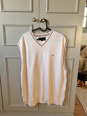 Vit väst från Tommy Hilfiger - Vit stickad väst från Tommy Hilfiger med v-ringning och tunna vinröda ränder längs krage och ärmöppningar. Klassisk logga broderad på bröstet. Perfekt för en preppy look och enkel att matcha med skjorta eller t-shirt.
