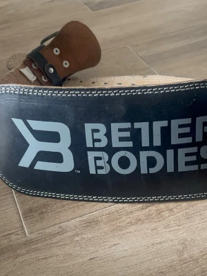 Svart lyftarbälte Better Bodies - Säljer ett svart lyftarbälte från Better Bodies med vit kontrastsöm och stor logga på framsidan. Bältet är gjort i kraftigt läder med mockainsida och flera hål för justering. Perfekt för gym och styrketräning.