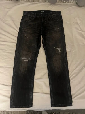 Svarta tapered jeans med slitningar - Säljer ett par svarta tapered jeans med coola slitningar och distressed detaljer på benen. Jeansen har klassisk femficksmodell och raka ben. Perfekta för dig som gillar en avslappnad men trendig stil.