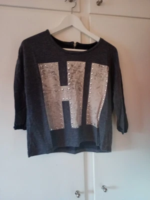 Mörkgrå topp med HI-tryck och nitar - Mörkgrå topp med stora bokstäverna HI i silverfärgat tyg framtill, dekorerad med små nitar runt bokstäverna. Trekvartsärm och avslappnad passform. Perfekt för en cool och avslappnad stil.