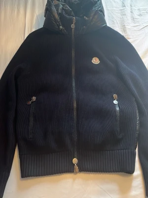 Svart pufferjacka från Moncler - Snygg svart cardigan från Moncler med stickade ärmar och quiltad baksida. Jackan har huva, två dragkedjefickor fram och ribbade muddar. Moncler-logga på bröstet och dragkedja hela vägen. Perfekt för allt. Den är storlek 2/M. Den har lite slitna dragkedjor men annars är den som helt ny. Den är köpt ifrån Merch Sweden. 