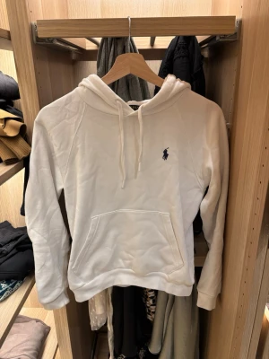 Vit hoodie från Polo Ralph Lauren - Snygg vit hoodie från Polo Ralph Lauren med klassisk huva och dragsko. Har en stor magficka och den ikoniska broderade loggan i mörkblått på bröstet. Tillverkad i mjukt material som känns skönt mot huden. Perfekt för en clean och stilren look. Ny pris c 2000 säljer för den inte kommer till användning