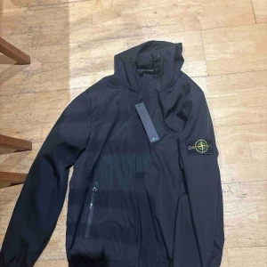 Svart vindjacka från Stone Island - Snygg svart vindjacka från Stone Island med huva och dragkedja framtill. Jackan har två sidofickor med dragkedja och den klassiska Stone Island-loggan på vänster ärm. Materialet är lätt och vindtätt, perfekt för blåsiga dagar.