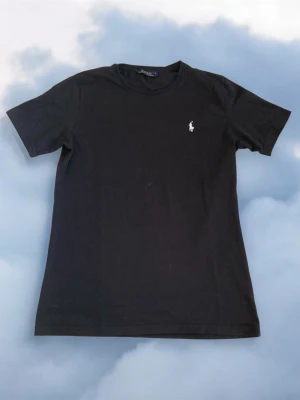 Svart t-shirt från Ralph Lauren - Klassisk svart t-shirt från Ralph Lauren med den ikoniska vita broderade loggan på bröstet. T-shirten har rund halsringning och korta ärmar. Tillverkad i mjuk bomull för en skön känsla och enkel stil.