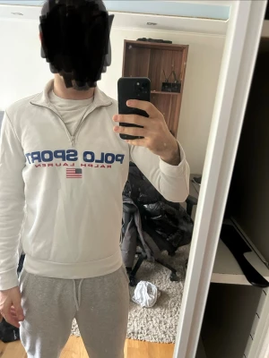 Vit quarter zip från Polo Sport - Säljer en vit quarter zip sweatshirt från Polo Sport Ralph Lauren. Tröjan har blå logga och amerikansk flagga på bröstet, hög krage med dragkedja och ribbade muddar. Perfekt för en sportig och avslappnad stil.