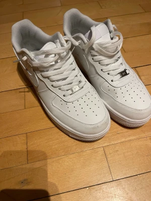 Nike Air Force 1 vita låga sneakers - Klassiska Nike Air Force 1 sneakers i helvitt läder med perforerad tåbox, vita skosnören och den ikoniska swoosh-loggan på sidan. Tjock sula med AIR-detalj och låg profil. Perfekta för dig som gillar stilrena och tidlösa sneakers.Pris går att diskuteras,använda ca 3 gånger