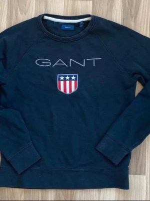Mörkblå sweatshirt från GANT - Mörkblå sweatshirt från GANT med broderad logga och sköld med stjärnor och ränder på bröstet. Klassisk rund halsringning och ribbade muddar vid ärmslut och nederkant. Perfekt för en avslappnad och stilren look.