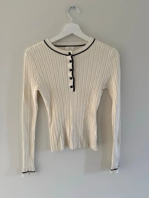 Vit ribbad tröja med svarta detaljer H&M - Vit långärmad ribbad tröja från H&M med svarta knappar och svarta kantdetaljer vid halsringning och ärmslut. Tröjan har en smal passform och är tillverkad i ett stretchigt material. Inprincip ny och storlek s.