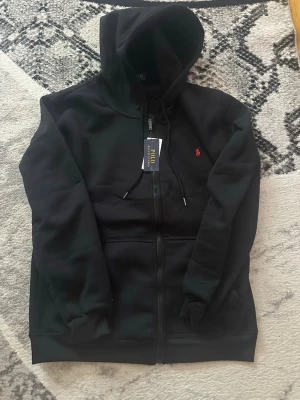 Svart hoodie från Polo Ralph Lauren - Svart hoodie med dragkedja från Polo Ralph Lauren. Klassisk design med huva och dragsko, samt fickor framtill. Liten röd broderad logga på bröstet ger en clean och stilren look. Perfekt för dig som gillar streetwear och premiumkänsla.