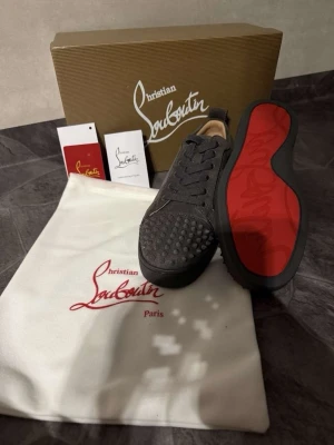 Christian Louboutin  - Snygga gråa sneakers från Christian Louboutin med klassisk röd sula och nitar på tån. Skorna har en låg profil, snörning och är tillverkade i mocka. Perfekta för dig som vill sticka ut med exklusiv design och detaljer.